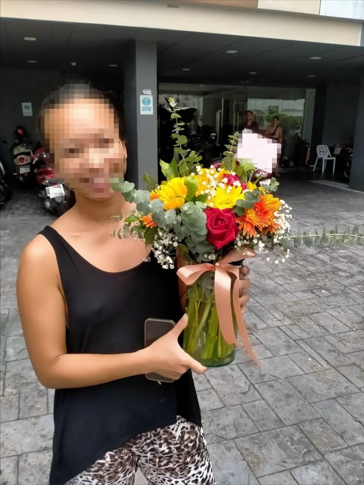 ลูกค้ากำลังรับแจกันดอกไม้ที่คล้ายกัน มีทานตะวันเหลือง เยอบีร่าส้ม กุหลาบแดง ยิปโซขาว (สินค้าอื่น)